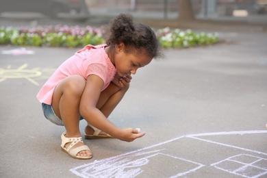 Little African-American child drawing house with chalk on asphalt