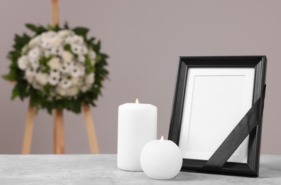 Photo frame with black ribbon, burning candles on table and wreath of plastic flowers near grey wall, space for text. Funeral attributes