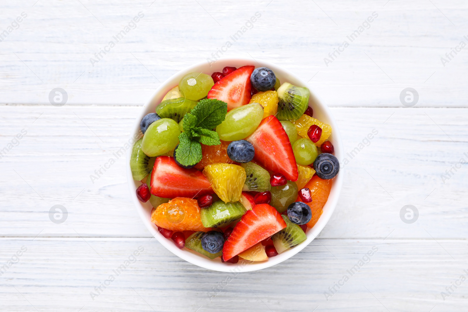 Photo of Delicious fresh fruit salad in bowl on white wooden table, top view