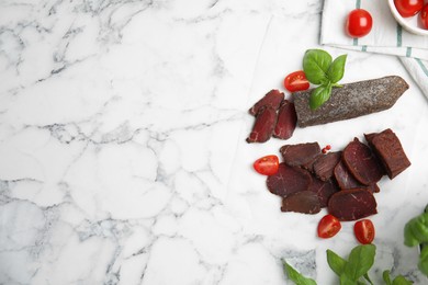 Photo of Delicious dry-cured beef basturma with basil and tomatoes on white marble table, flat lay. Space for text