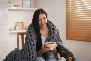 Woman with chunky knit blanket and cup in armchair at home