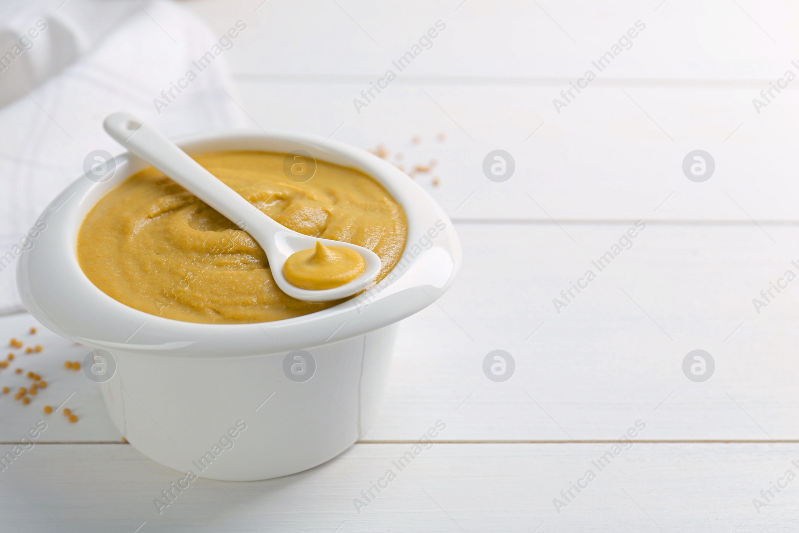 Photo of Spoon and bowl of tasty mustard sauce on white wooden table, space for text