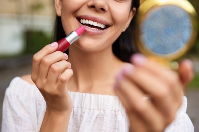 Photo of Woman with cosmetic pocket mirror applying lipstick outdoors, closeup