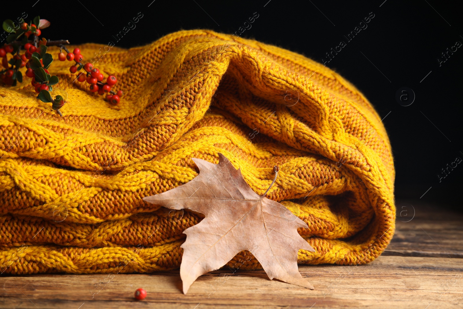 Photo of Orange knitted plaid with red berries and dry leaf on wooden table, closeup