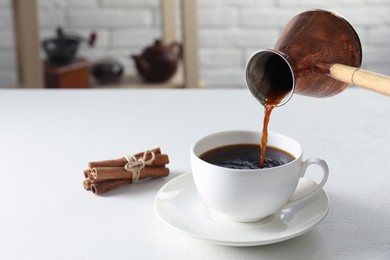 Turkish coffee. Pouring brewed beverage from cezve into cup at white table indoors