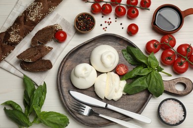 Delicious burrata cheese with basil and tomatoes served on white wooden table, flat lay