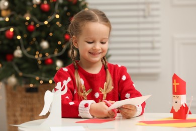 Photo of Cute little girl cutting paper at table with Saint Nicholas toy indoors