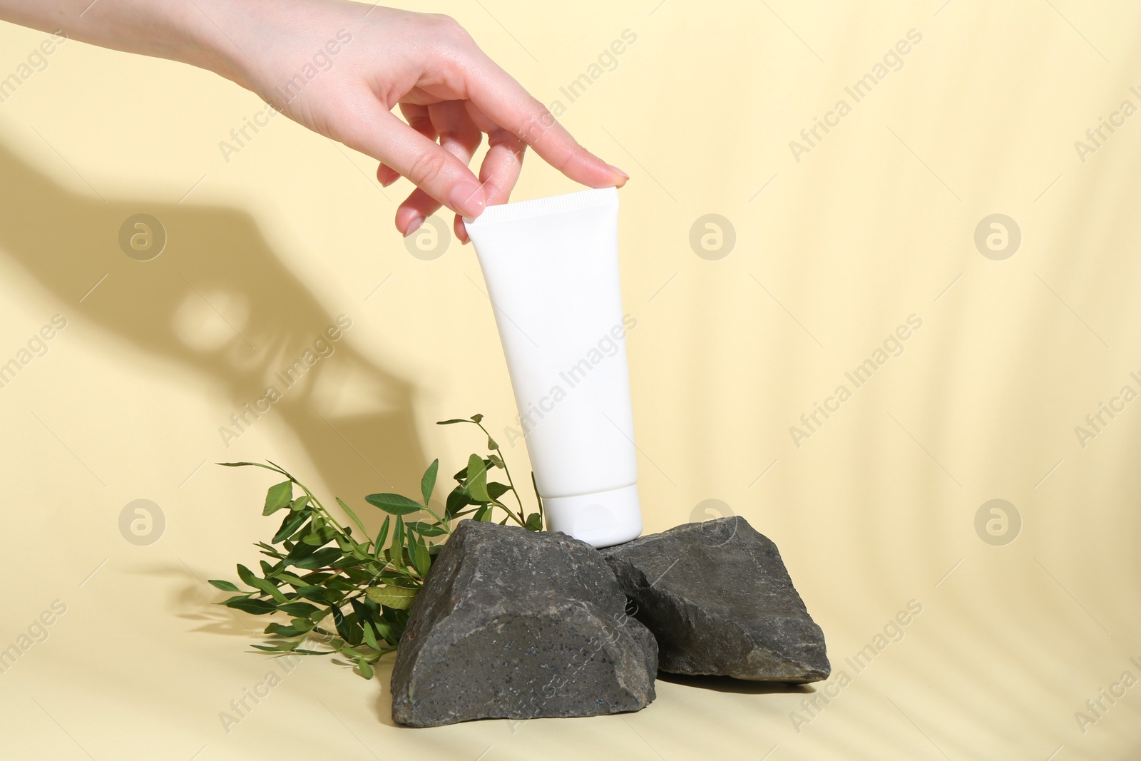 Photo of Woman with tube of cream, branches and stones on light yellow background, closeup