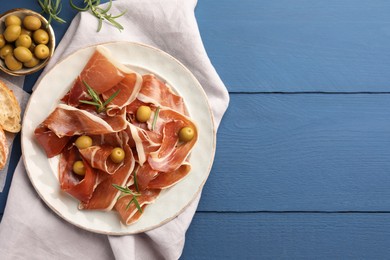 Slices of tasty cured ham, rosemary and olives on blue wooden table, flat lay. Space for text