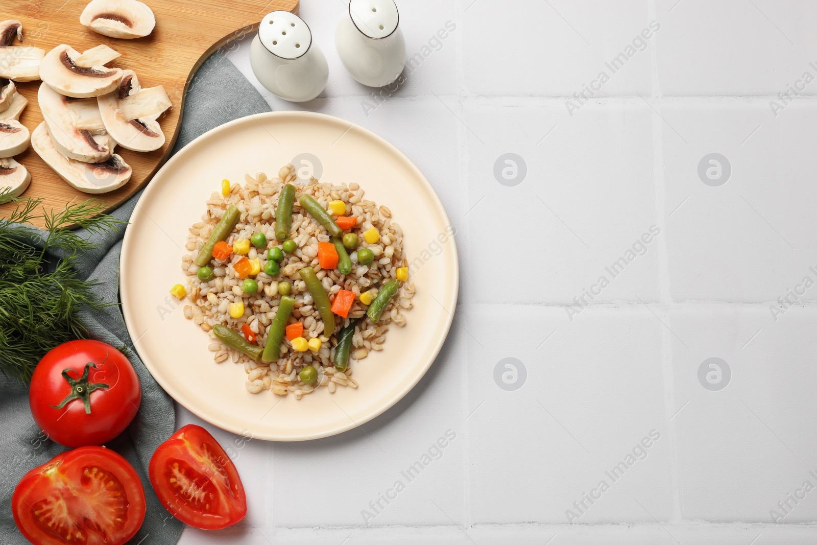 Photo of Delicious pearl barley with vegetables served on white tiled table, flat lay. Space for text