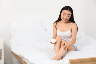 Beautiful young Asian woman applying body cream on leg in bedroom, space for text