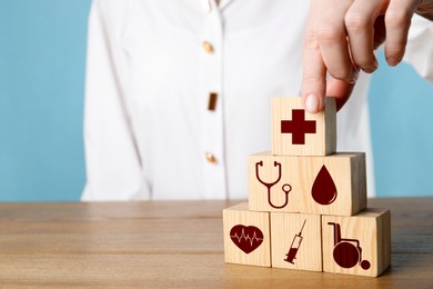 Woman building pyramid of cubes with different icons on wooden table against light blue background, closeup. Insurance concept