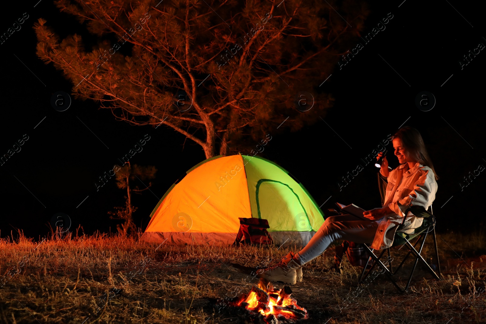 Photo of Young woman with flashlight reading book near bonfire at night. Camping season