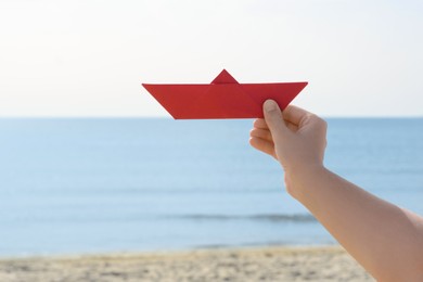 Child holding red paper boat near sea, closeup