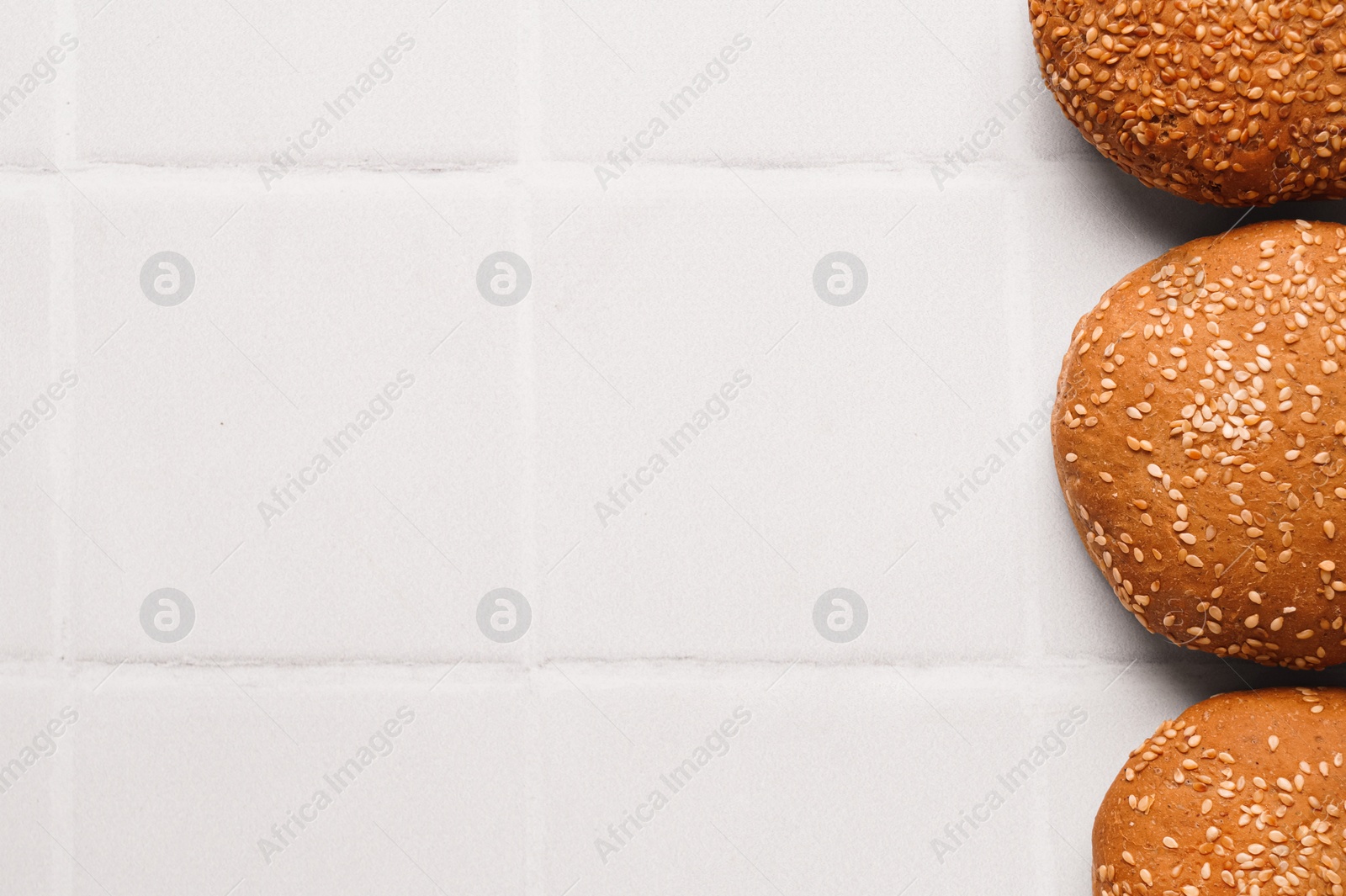 Photo of Fresh buns with sesame seeds on white tiled table, flat lay. Space for text