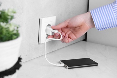 Photo of Woman plugging power bank into socket at white table indoors, closeup