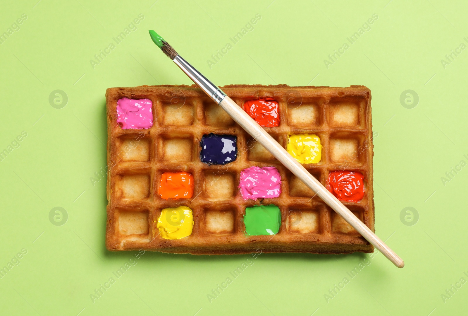 Photo of Waffle with different paints and brush on light green background, top view