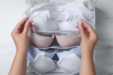 Woman putting bra into organizer with underwear at white marble table, top view