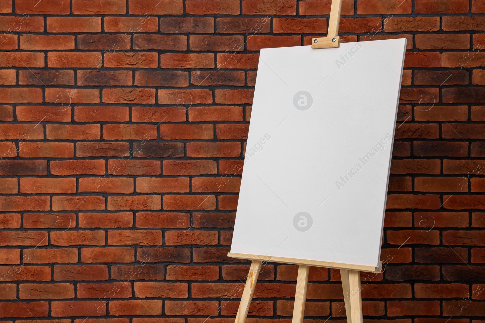Photo of Wooden easel with blank canvas near brick wall. Space for text