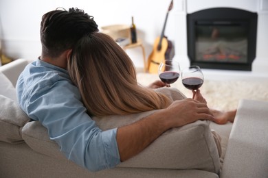 Photo of Couple with glasses of wine resting on sofa near fireplace at home, back view