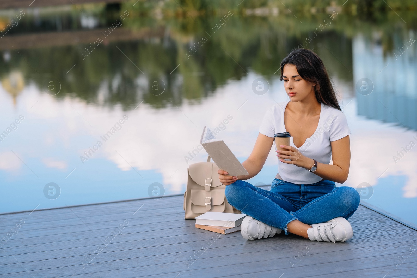 Photo of Young woman with cup of coffee reading book on pier near lake