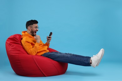 Photo of Happy young man using smartphone on bean bag chair against light blue background. Space for text