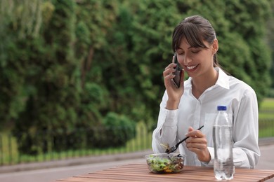 Photo of Happy businesswoman talking on smartphone while having lunch at wooden table outdoors, space for text