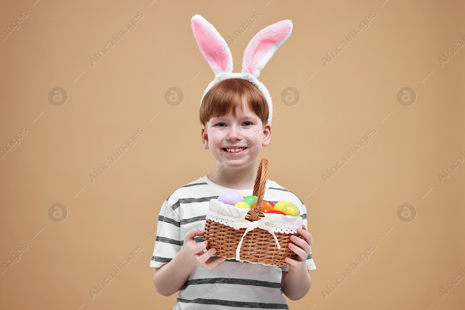Photo of Easter celebration. Cute little boy with bunny ears and wicker basket full of painted eggs on dark beige background