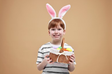 Photo of Easter celebration. Cute little boy with bunny ears and wicker basket full of painted eggs on dark beige background