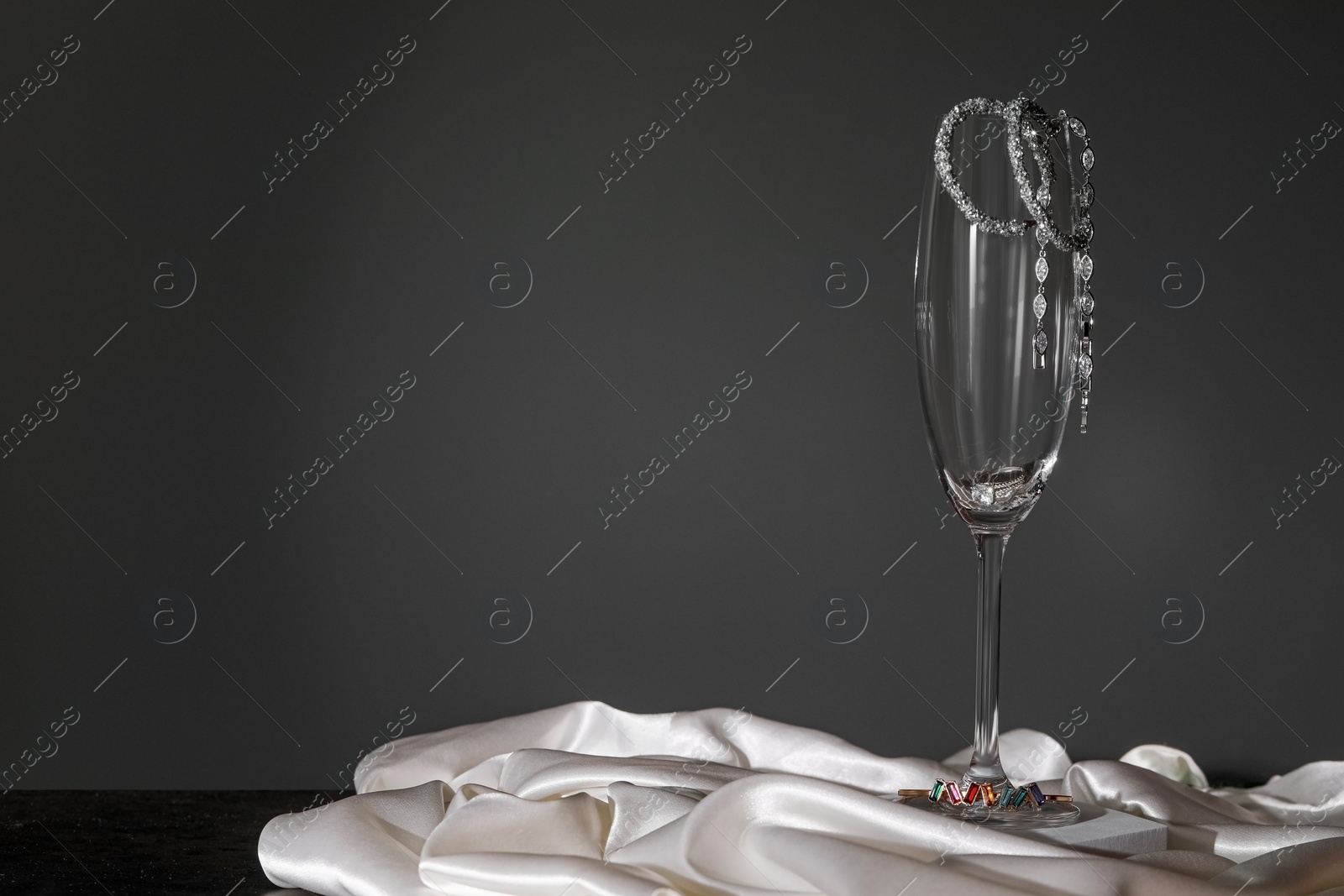 Photo of Elegant jewelry. Glass with luxury earrings on white silk against black background. Space for text