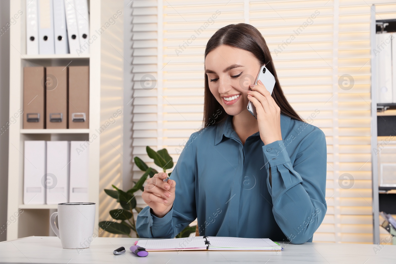 Photo of Happy woman taking notes while talking on smartphone at white marble table in office