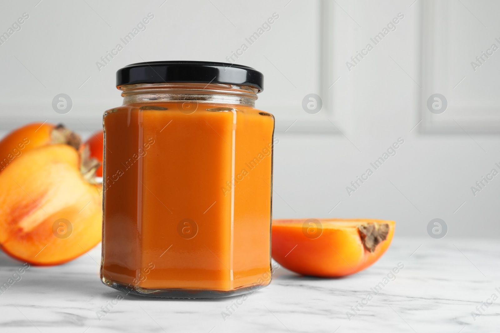 Photo of Delicious persimmon jam and fresh fruits on white marble table. Space for text