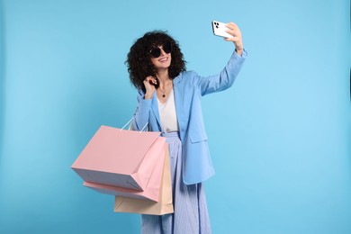 Happy young woman with shopping bags taking selfie on light blue background