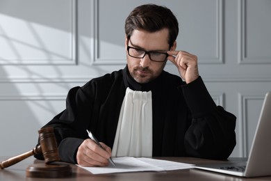Judge with gavel and laptop writing in papers at wooden table indoors