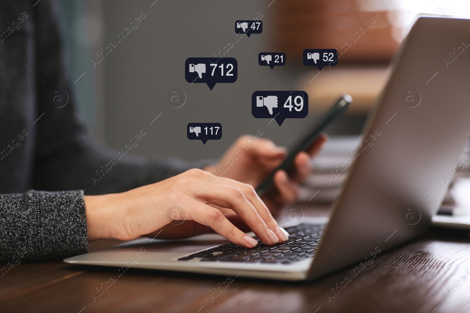 Image of Closeup view of young woman using modern laptop with smartphone indoors and virtual dislike icons at table. Cyberbullying concept