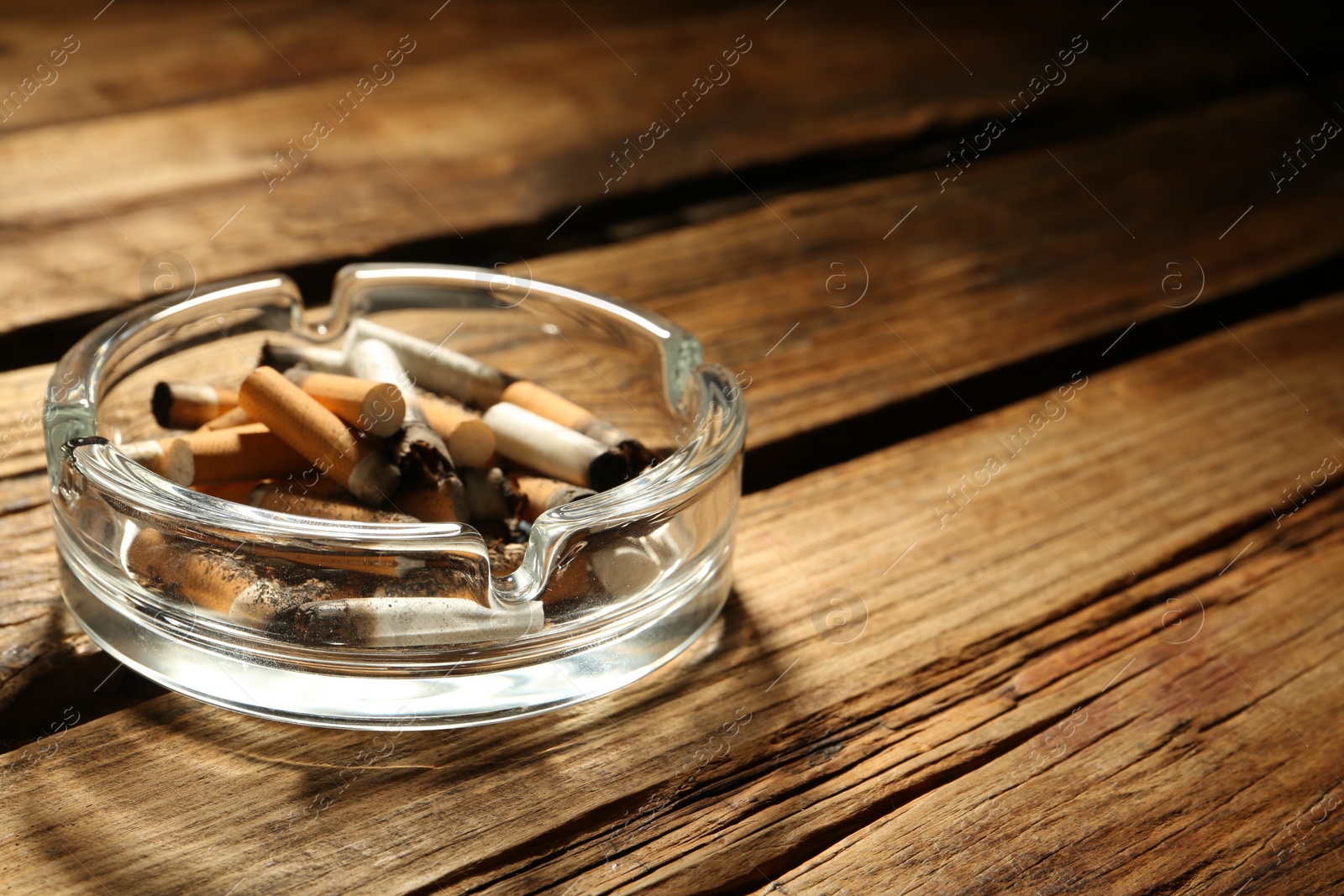 Photo of Glass ashtray with cigarette stubs on wooden table, closeup. Space for text