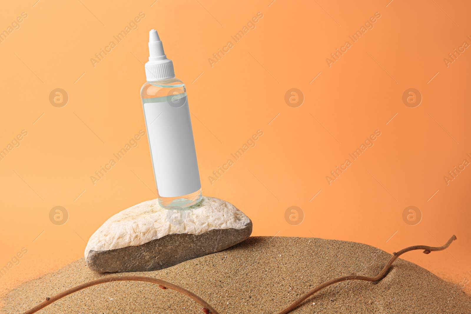 Photo of Bottle with serum, branch and stone on sand against orange background, space for text. Cosmetic product