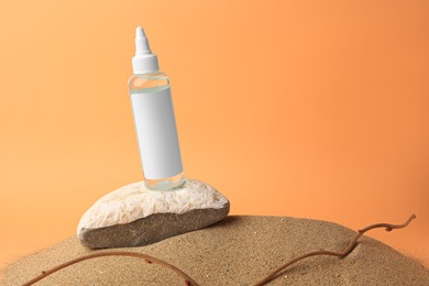 Photo of Bottle with serum, branch and stone on sand against orange background, space for text. Cosmetic product