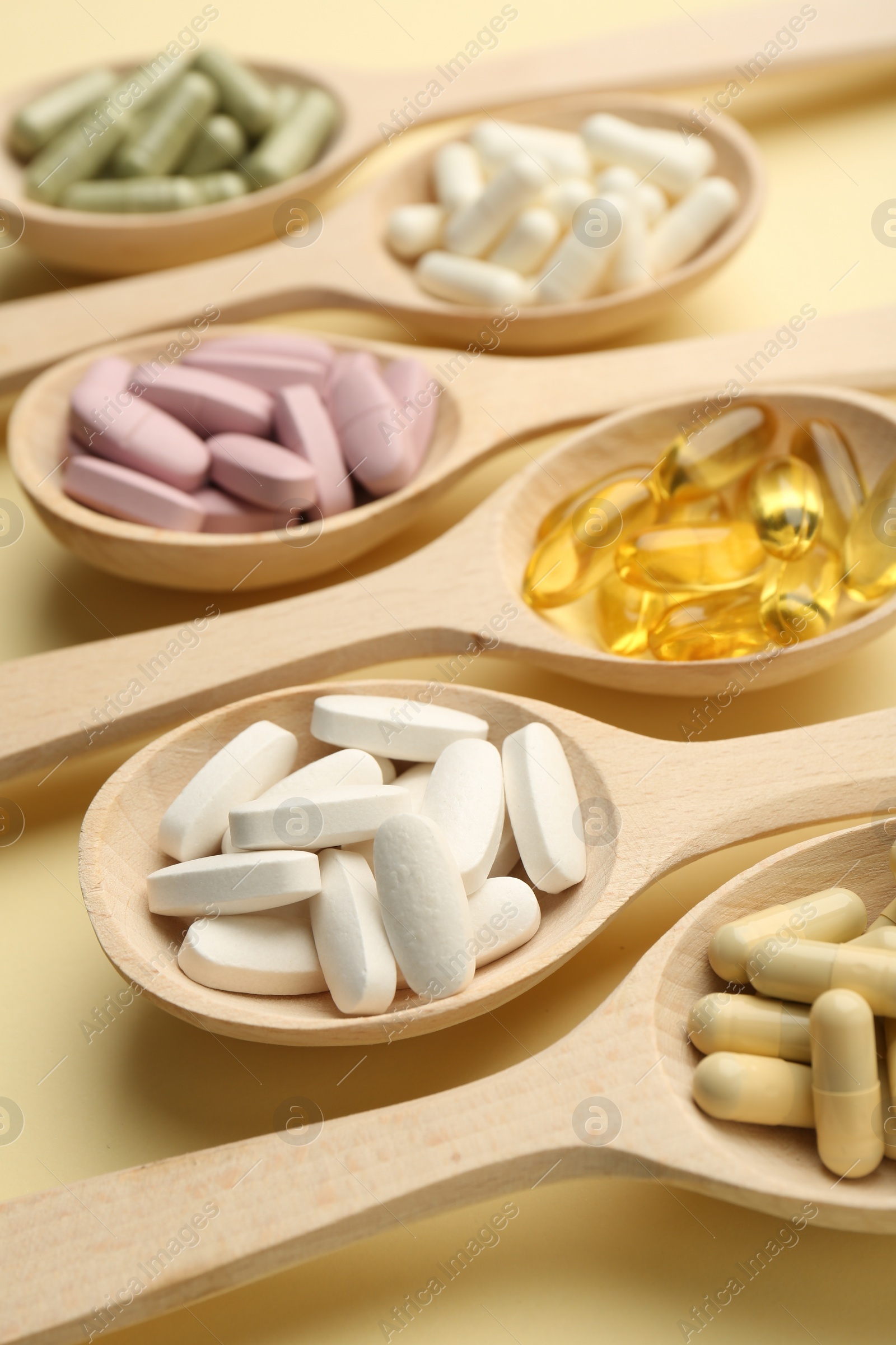 Photo of Different vitamin pills in spoons on pale yellow background, closeup