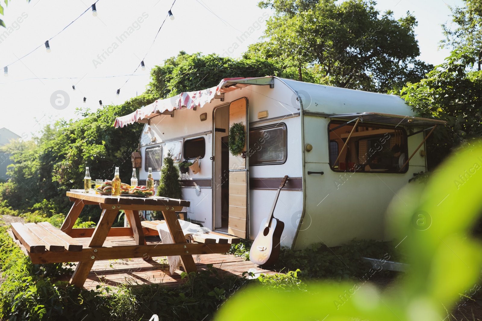 Photo of Wooden table with food and bottles of beer near trailer on sunny day. Camping season