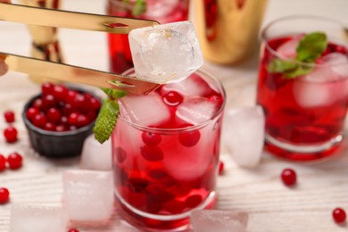 Putting ice cube into glass with tasty cranberry cocktail at light table, closeup