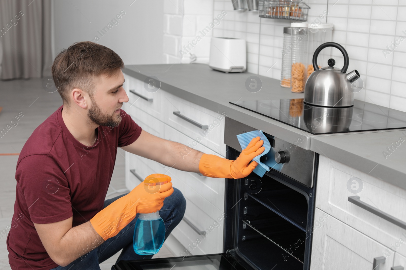 Photo of Young man cleaning oven with rag and detergent in kitchen