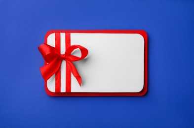 Photo of Blank gift card with red bow on blue background, top view