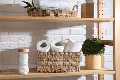 Photo of Toilet paper rolls in wicker basket, floral decor and cotton pads on wooden shelf near white brick wall
