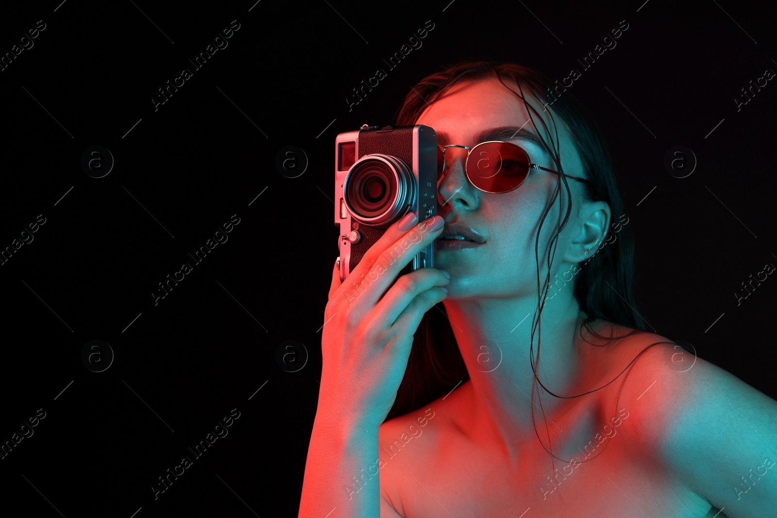 Photo of Beautiful woman with sunglasses and vintage camera posing in neon lights against black background. Space for text