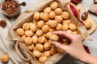 Woman taking delicious nut shaped cookie with boiled condensed milk from wicker basket at light wooden table, top view