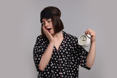 Photo of Tired young woman with sleep mask and alarm clock yawning on light grey background. Insomnia problem