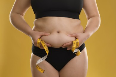 Woman with measuring tape touching belly fat on goldenrod background, closeup. Overweight problem