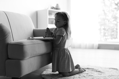 Photo of Cute little girl with beads praying over Bible in living room, black and white effect. Space for text