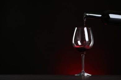 Photo of Pouring wine from bottle into glass on table against dark background, space for text